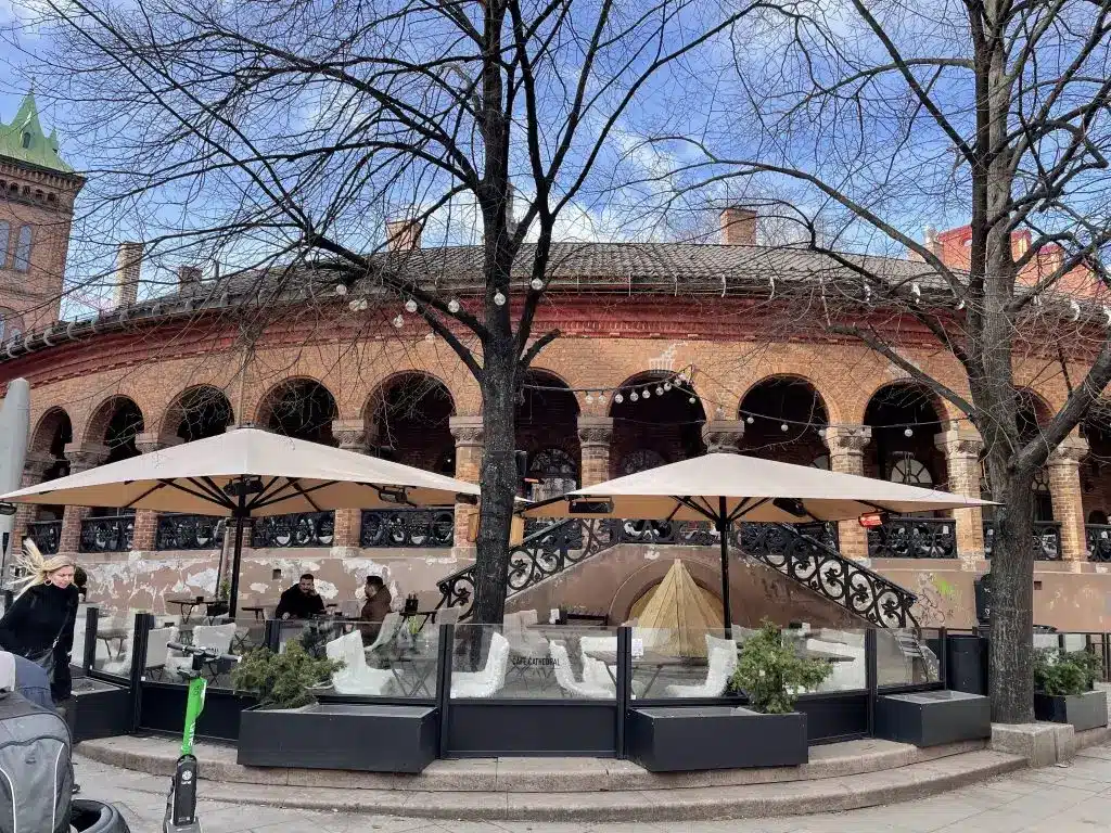 Ein charmantes Außenrestaurant mit mehreren Sonnenschirmen und Sitzbereichen, eingefasst von hohen, transparenten Windschutzscheiben, vor dem Hintergrund eines historischen Backsteingebäudes mit Bogenfenstern und dekorativen Verzierungen. Kahl wirkende Bäume stehen vor dem Restaurant, das unter einem klaren blauen Himmel an einem belebten städtischen Platz gelegen ist.